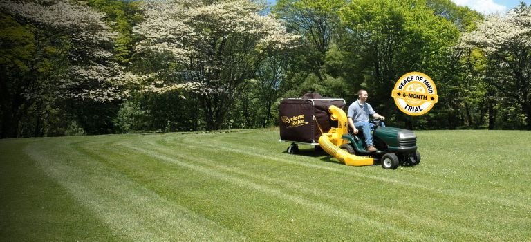 The Cyclone Rake lawn and leaf pickup machine being towed by a lawn tractor in a yard in spring
