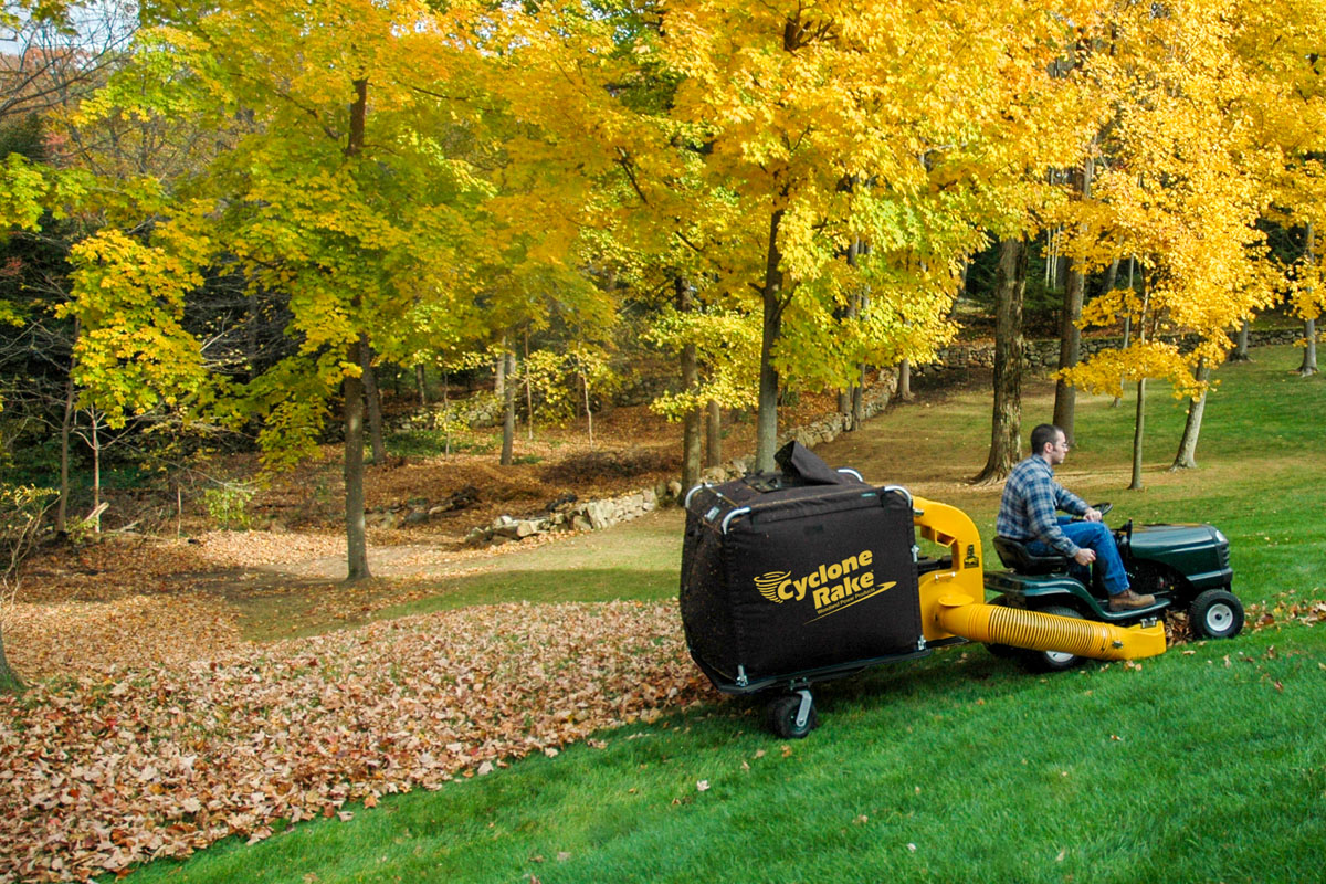 Cyclone Rake XL Super behind a lawn tractor mid-pass, lifting leaves from a wooded yard in fall