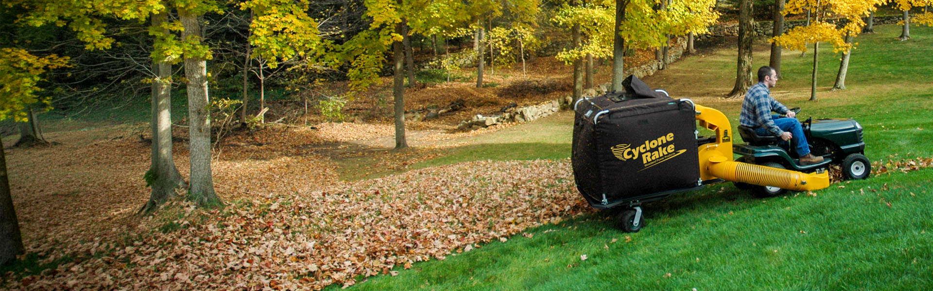 Cyclone Rake XL Super behind a lawn tractor mid-pass, lifting leaves from a wooded yard in fall